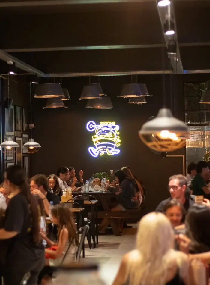 El salón de Sistons de noche, con el cartel de neón de la hamburguesa encendido sobre la pared negra y las mesas ocupadas.