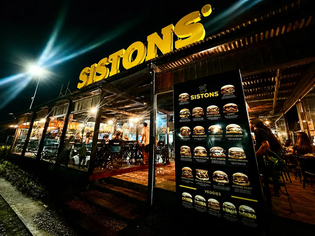 Frente del local de Sistons Burgers de noche, con el letrero encendido y el salón lleno.